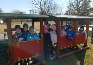 Grupa dzieci stoi w pociągu na placu zabaw, chłopiec trzyma maskotkę krasnala Hałabały.